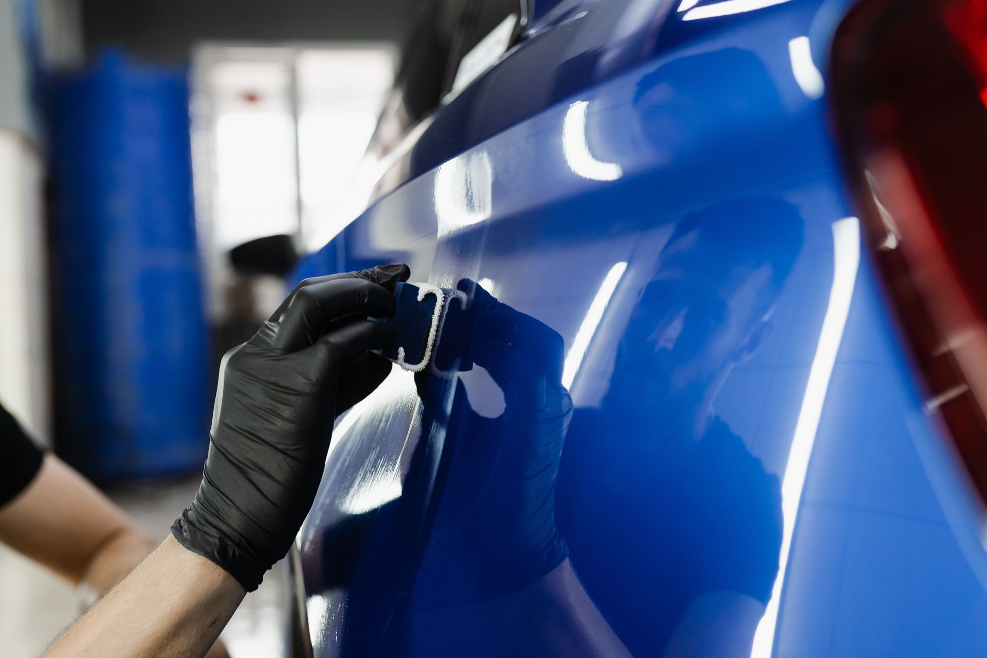 Gloved hand applying a clear coat to a shiny blue car door in a workshop