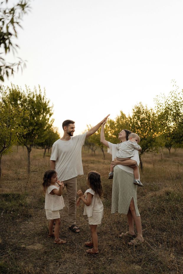Family in a sunlit orchard, with parents lifting children amid trees at sunset.