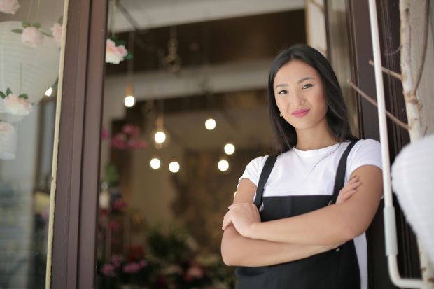 Woman in a white shirt and black apron standing with arms crossed in a doorway, with blurred lights behind her.
