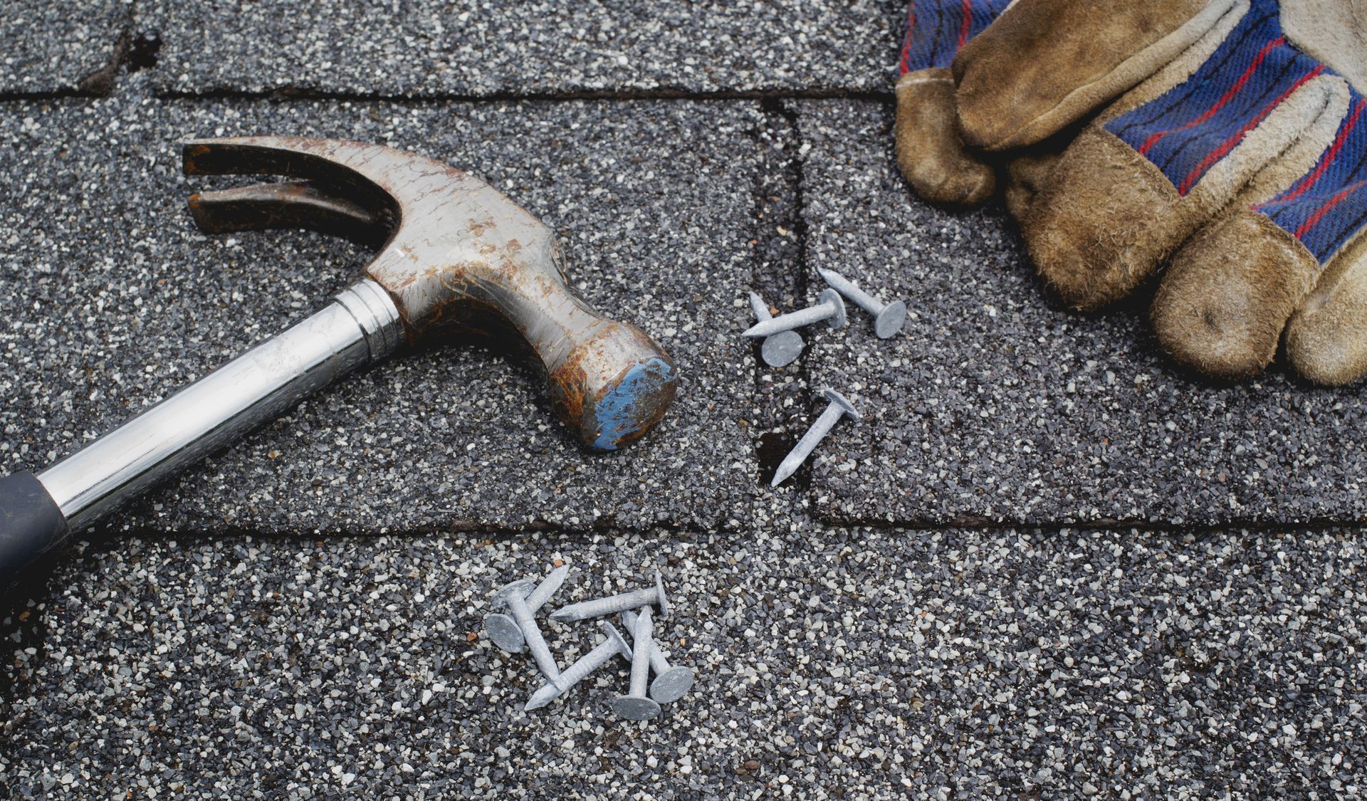 Hammer, nails, and work gloves on a dark gray asphalt shingle roof.