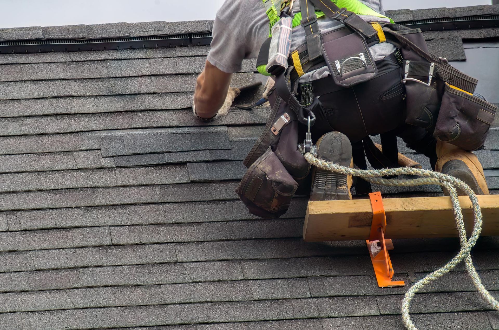 Roofer on a gray shingled roof, secured by a safety harness and rope.