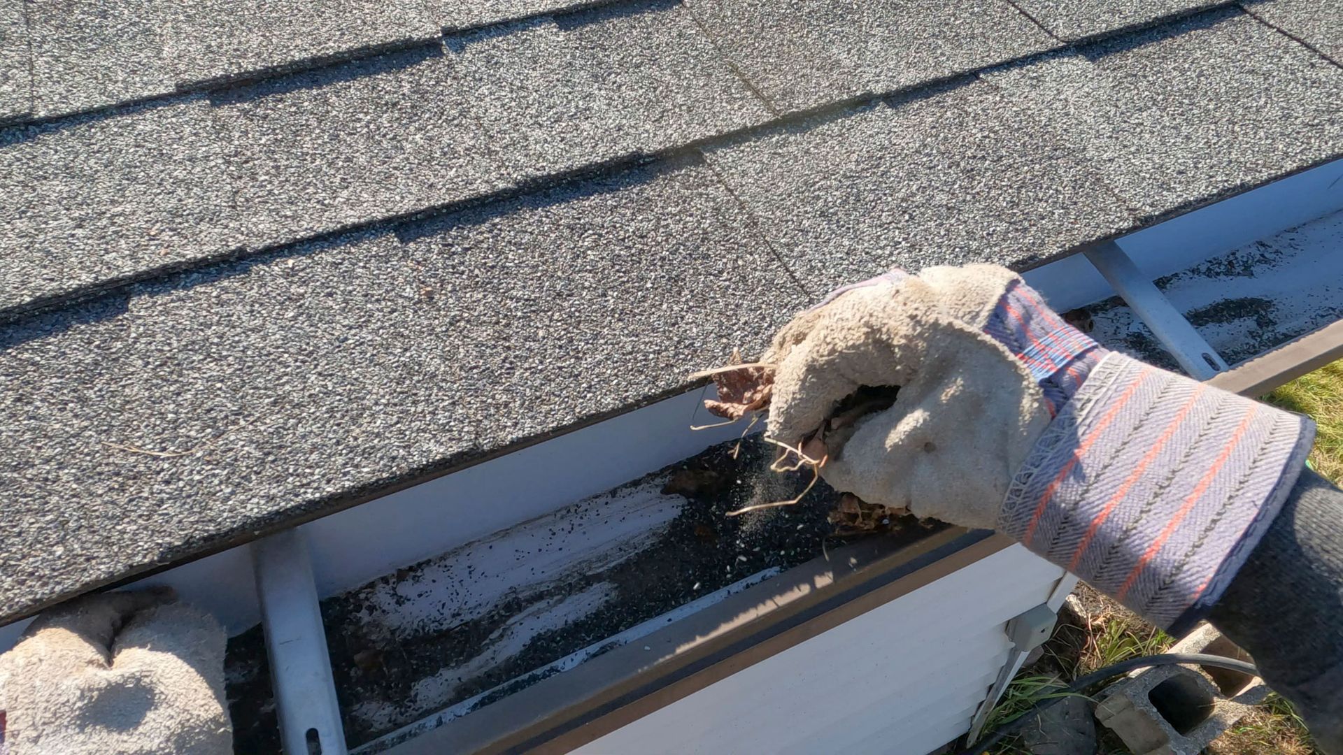 Person wearing gloves cleans a debris-filled gutter on a roof.