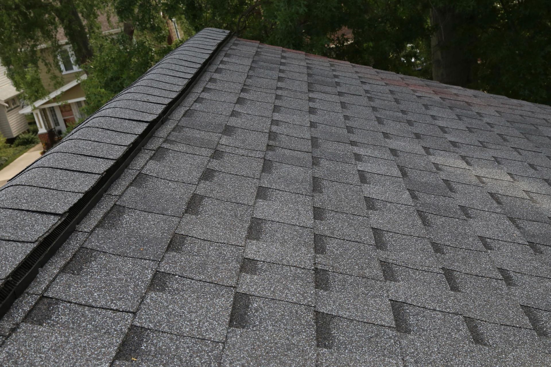 Gray asphalt shingle roof, close-up view. The roof is partially visible against a background of trees and houses.