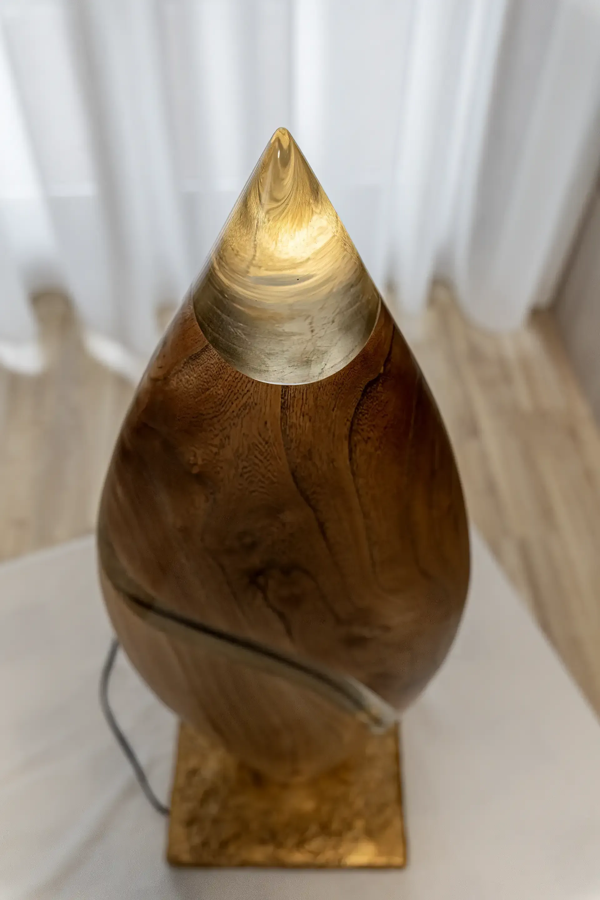 Lampada scultura in resina epossidica trasparente e legno. L'opera ha una forma a goccia con una sezione principale in legno e un inserto curvilineo in resina. La punta superiore è in resina trasparente dove viene emessa la luce. Opera di Guerino Rusconi.