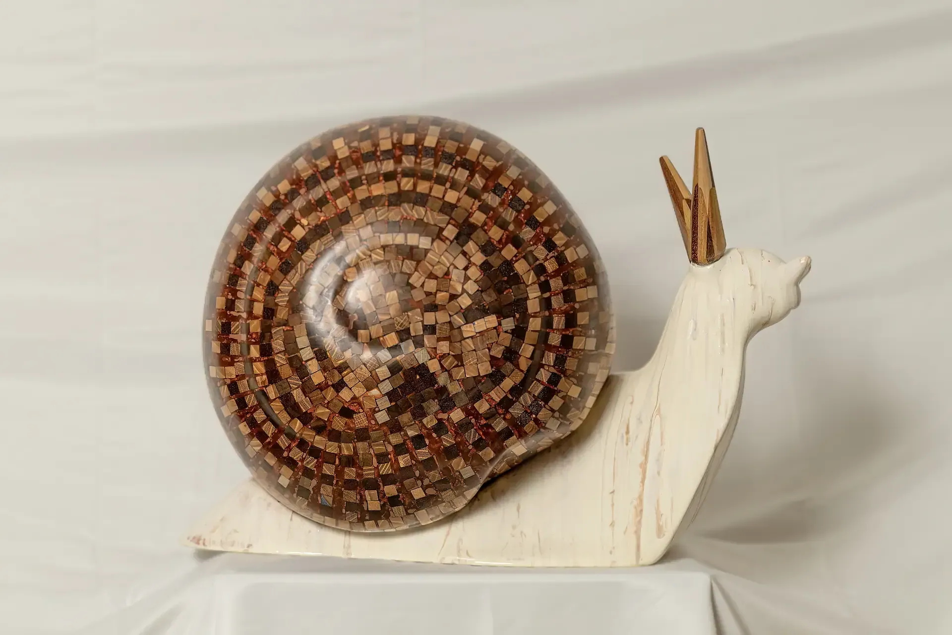Scultura di una chiocciola con corpo bianco di gesso ceramico e conchiglia marrone a mosaico di tasselli di legno rivestiti con resina trasparente. Inquadratura laterale che evidenzia il corpo e il legno venato e il guscio sferico a mosaico.