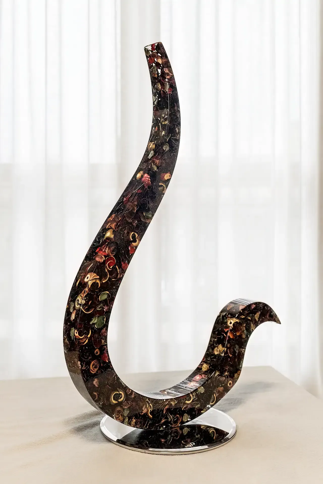 Scultura in resina epossidica con inclusioni di fiori essiccati multicolori, raffigurante una forma organica allungata. L' opera poggia su una base-piedistallo circolare in acciaio. Opera di Guerino Rusconi.