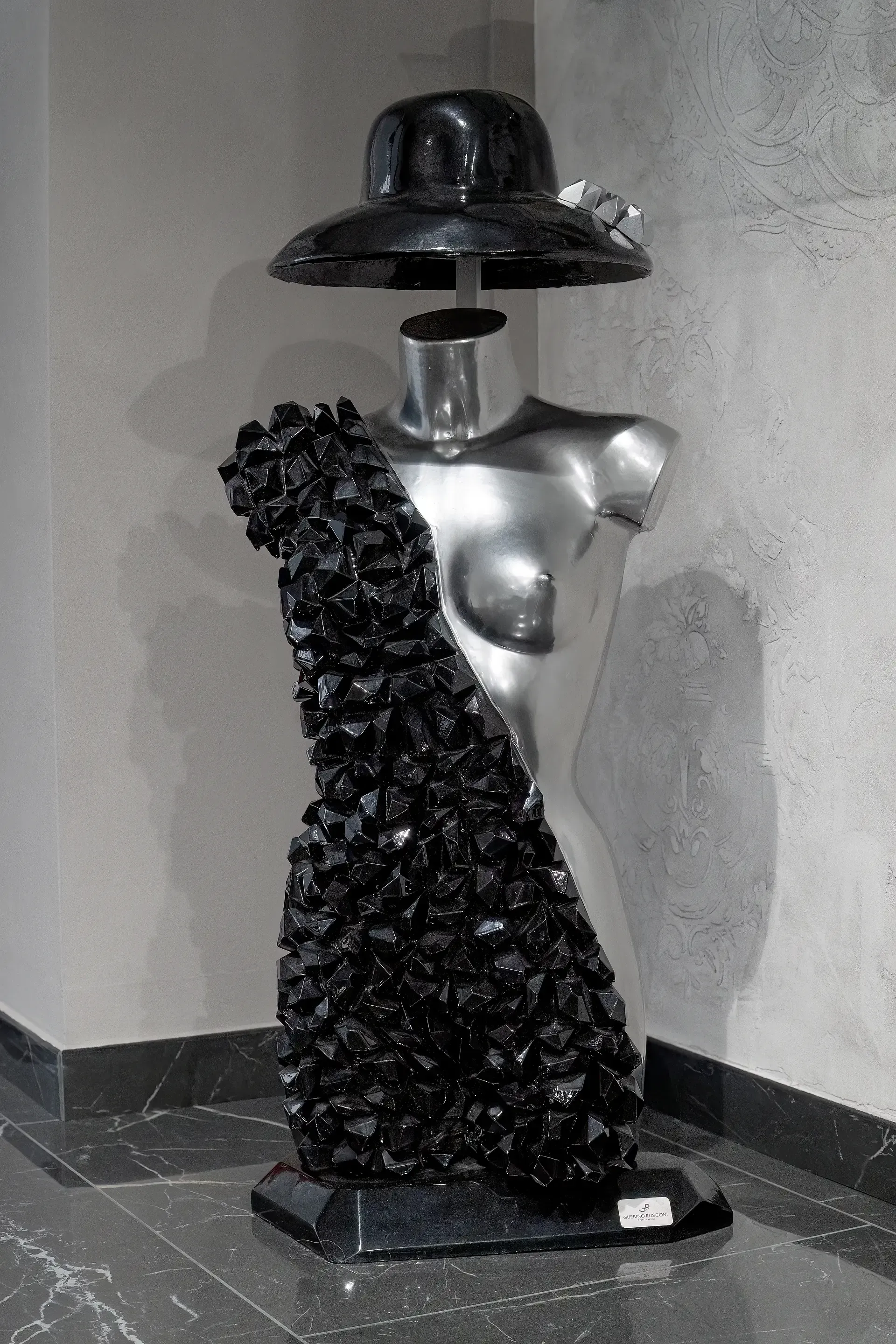 Scultura in resina e gesso ceramico. L'opera, realizzata dall'artista Guerino Rusconi, raffigura un busto femminile color argento. Sopra il busto troviamo un cappello a tesa larga in resina nera lucida in sospensione. La parte frontale presenta una struttura tridimensionale in resina nera, composta da elementi geometrici irregolari che ricorda la forma di un vestito.