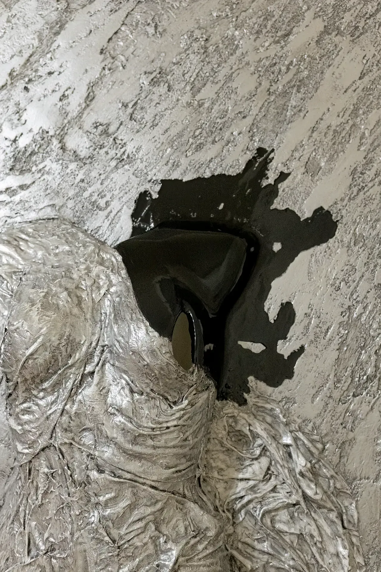 Dettaglio ravvicinato della scultura tridimensionale da parete con tecnica mista. L'opera presenta un fondo in tela con texture materica nei toni del grigio. In primo piano, si notano il drappeggio in materiale argenteo una inclusione di resina nera. Opera di Guerino Rusconi