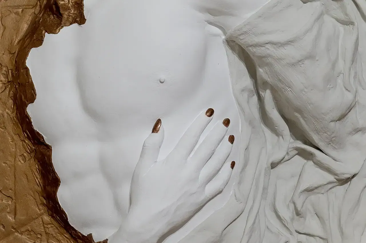 Dettaglio della scultura tridimensionale da parete con tecnica mista di torso di uomo. In primo piano, si notano la texture liscia del torso e le pieghe del drappeggio. Una mano in gesso ceramico, anch'essa di colore bianco, è appoggiata sul petto. Le unghie sono dipinte di un colore oro. Sullo sfondo, si intravede una superficie con texture materica nei toni del marrone e dell'oro. Opera di Guerino Rusconi.