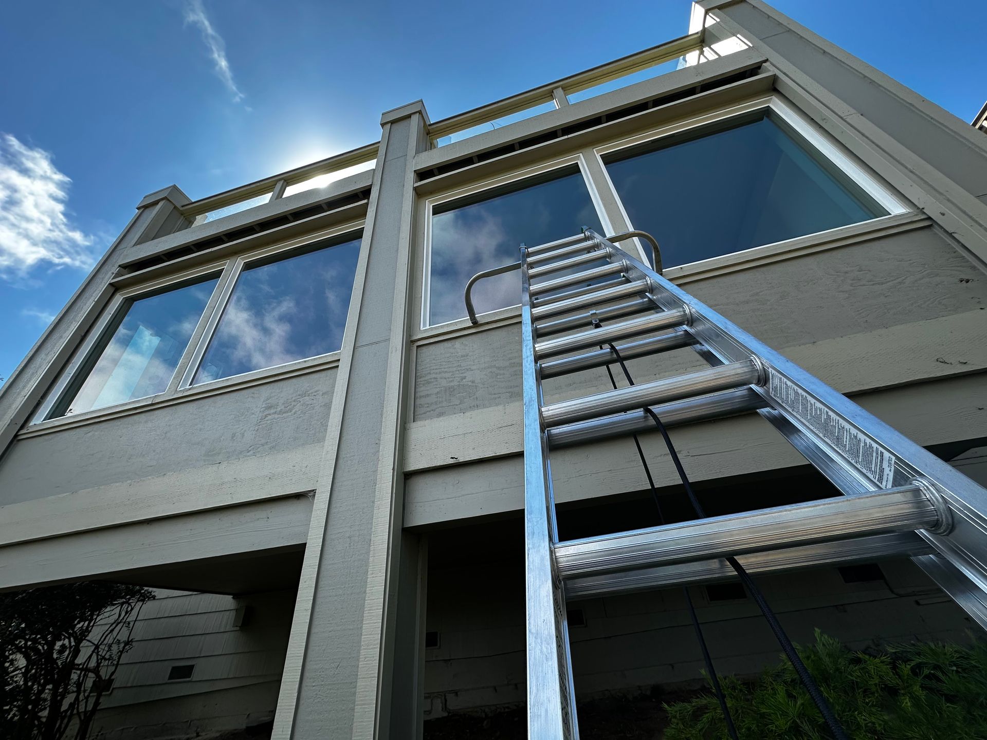 A silver ladder leans against a light-colored building, reaching towards the windows, under a blue sky.