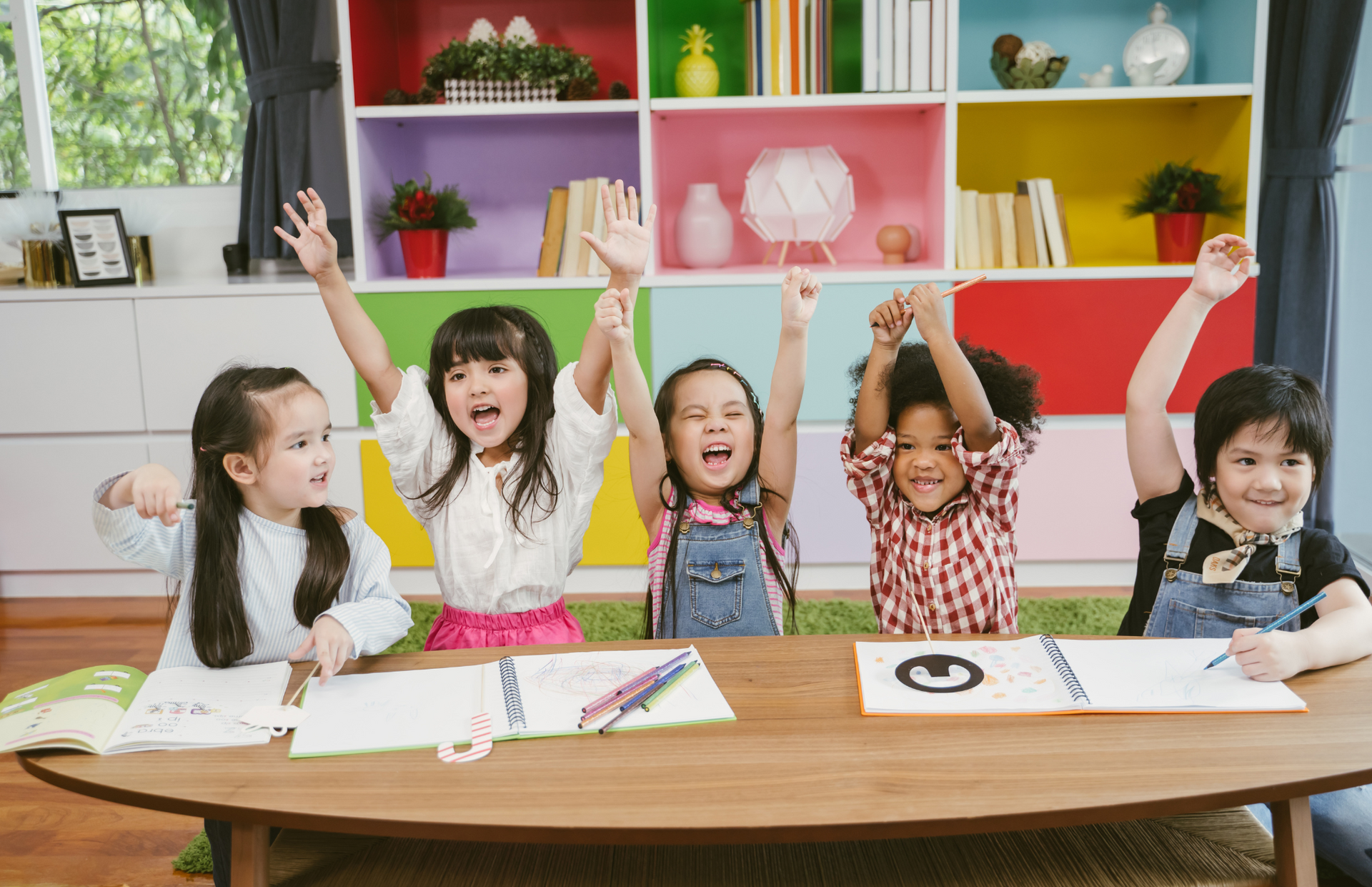 A group of children are sitting at a table with their hands in the air.