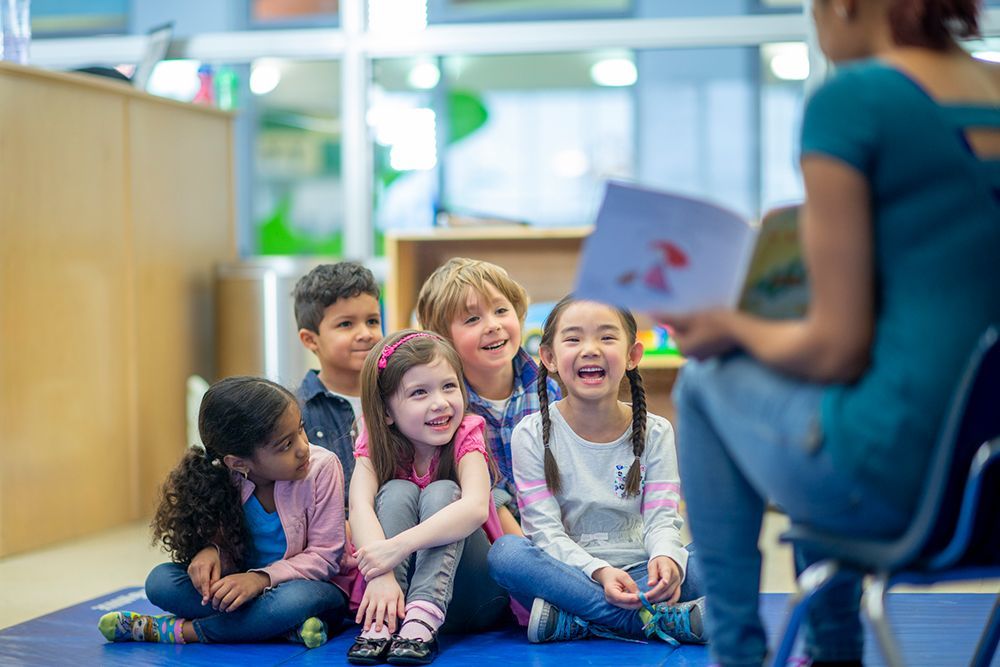 A group of children are sitting on the floor listening to a teacher read a book.