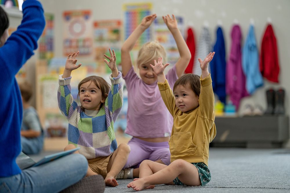 A group of children are sitting on the floor with their hands in the air.