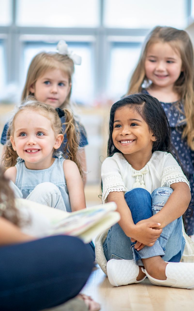 Four young children smiling and listening to someone reading in a classroom setting. Four young children smiling and listening to someone reading in a classroom setting.
