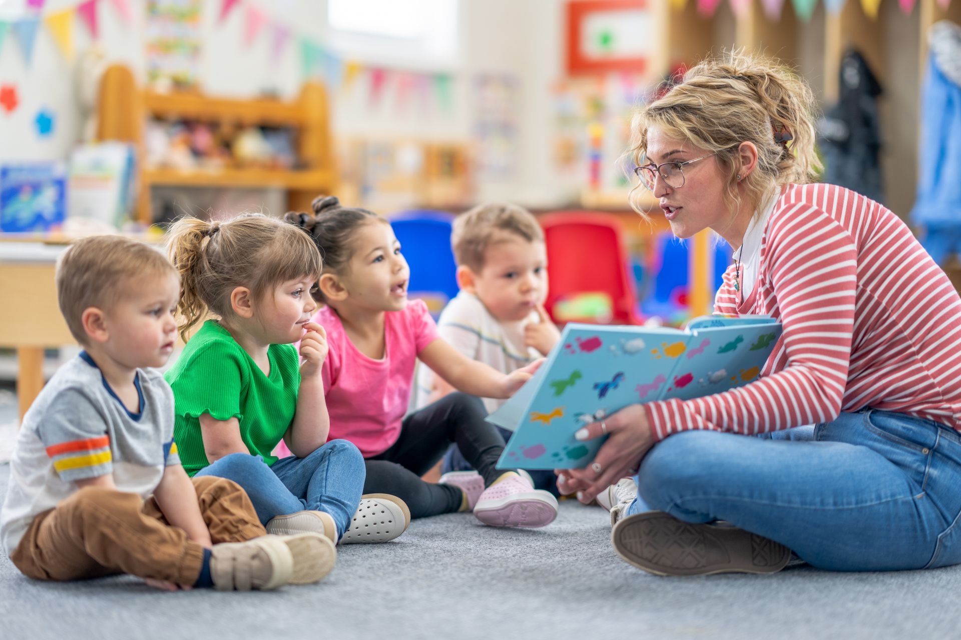 A teacher sitting on the floor, showing a book to a group of childs, highlighting a daycare center.