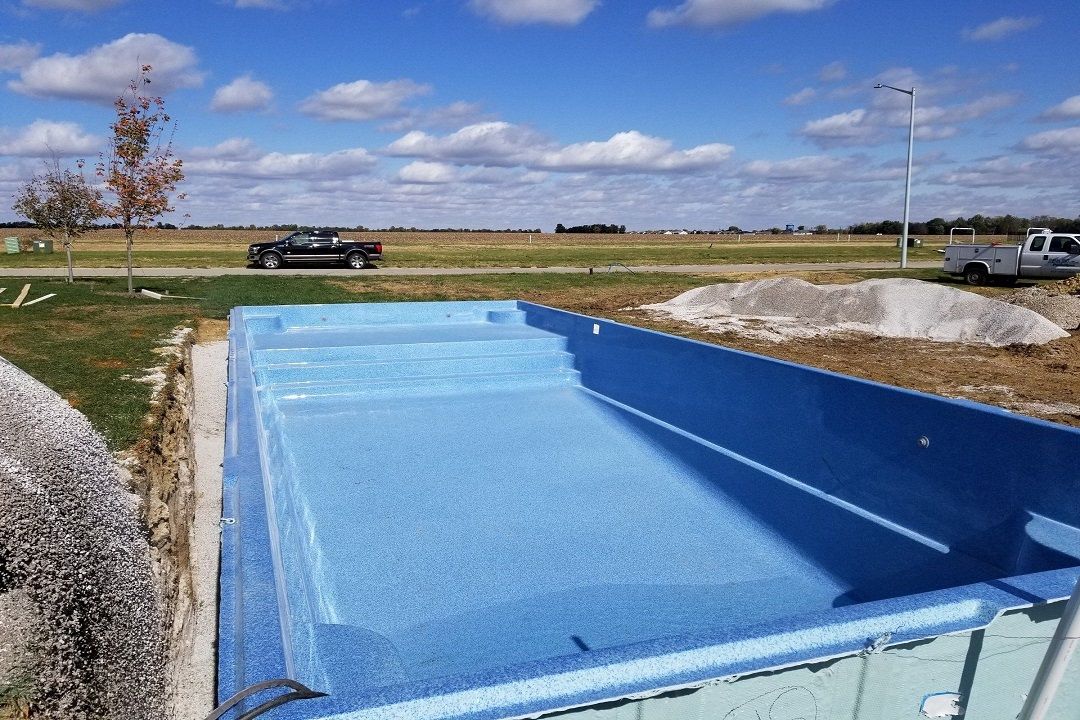 Blue pool liner installed in a rectangular pool excavation, with a car and construction equipment in the background.
