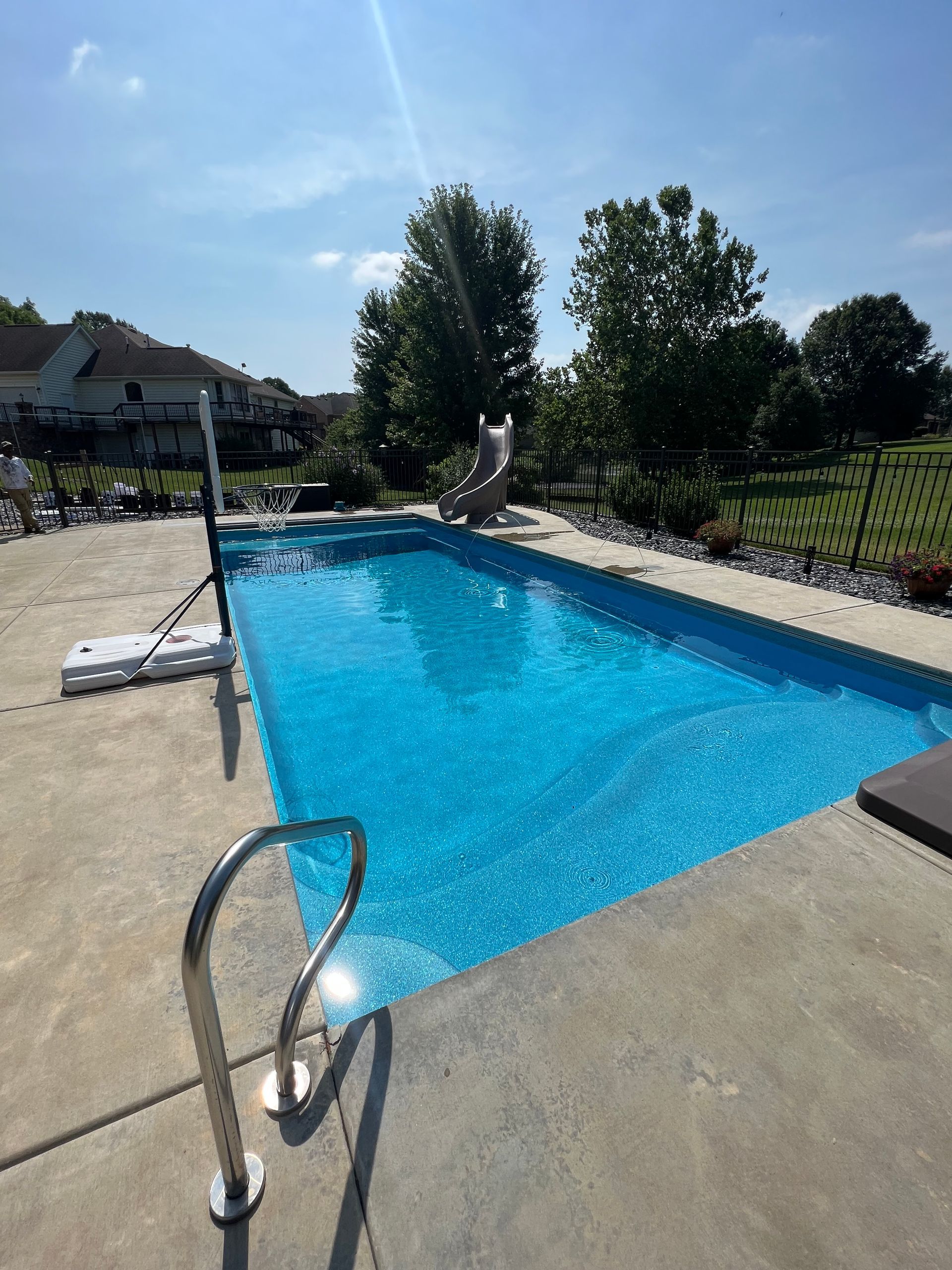 A rectangular blue swimming pool surrounded by concrete on a sunny day. A metal ladder is visible.