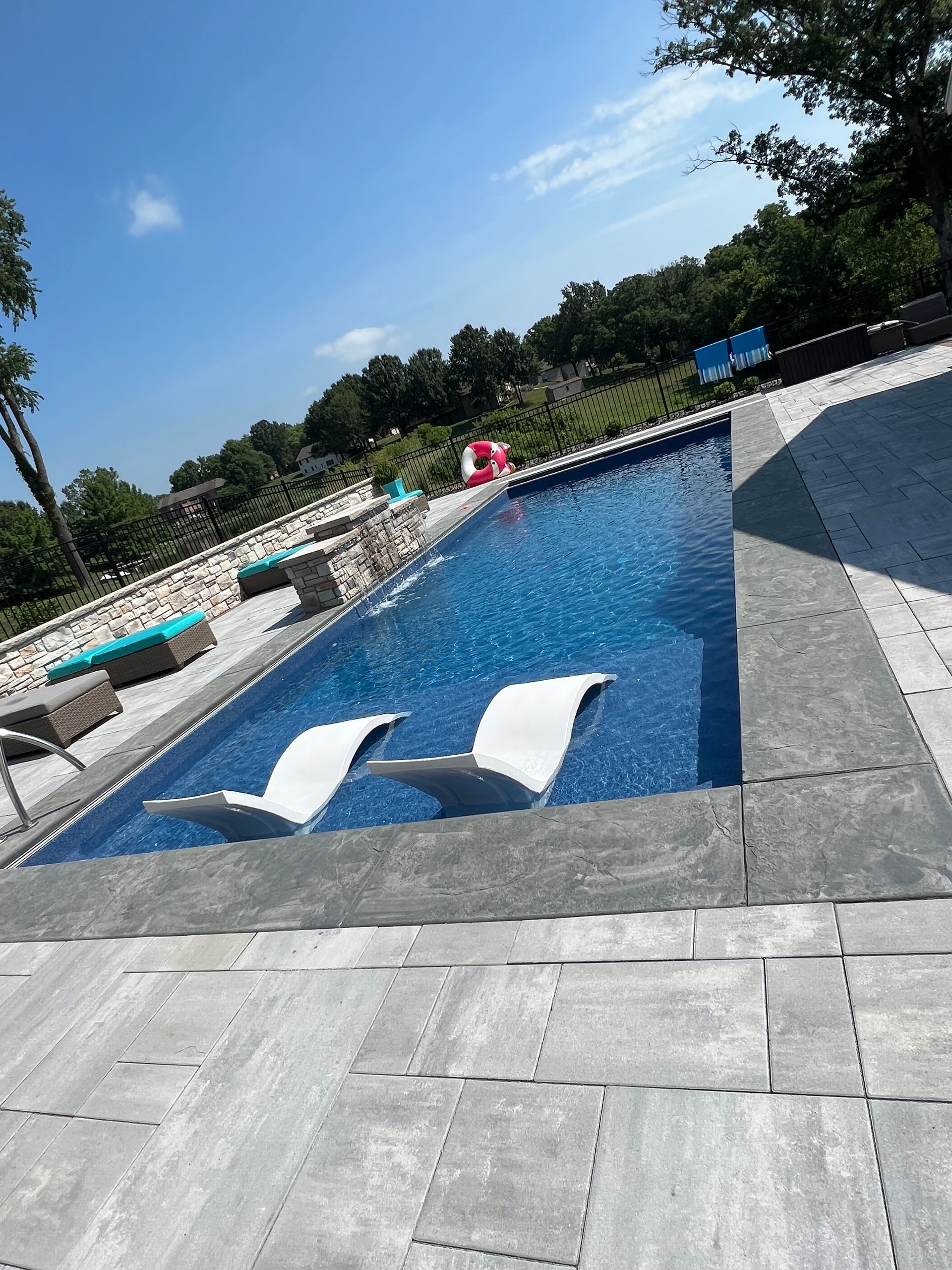 Swimming pool with two white lounge chairs in the water, surrounded by gray patio.