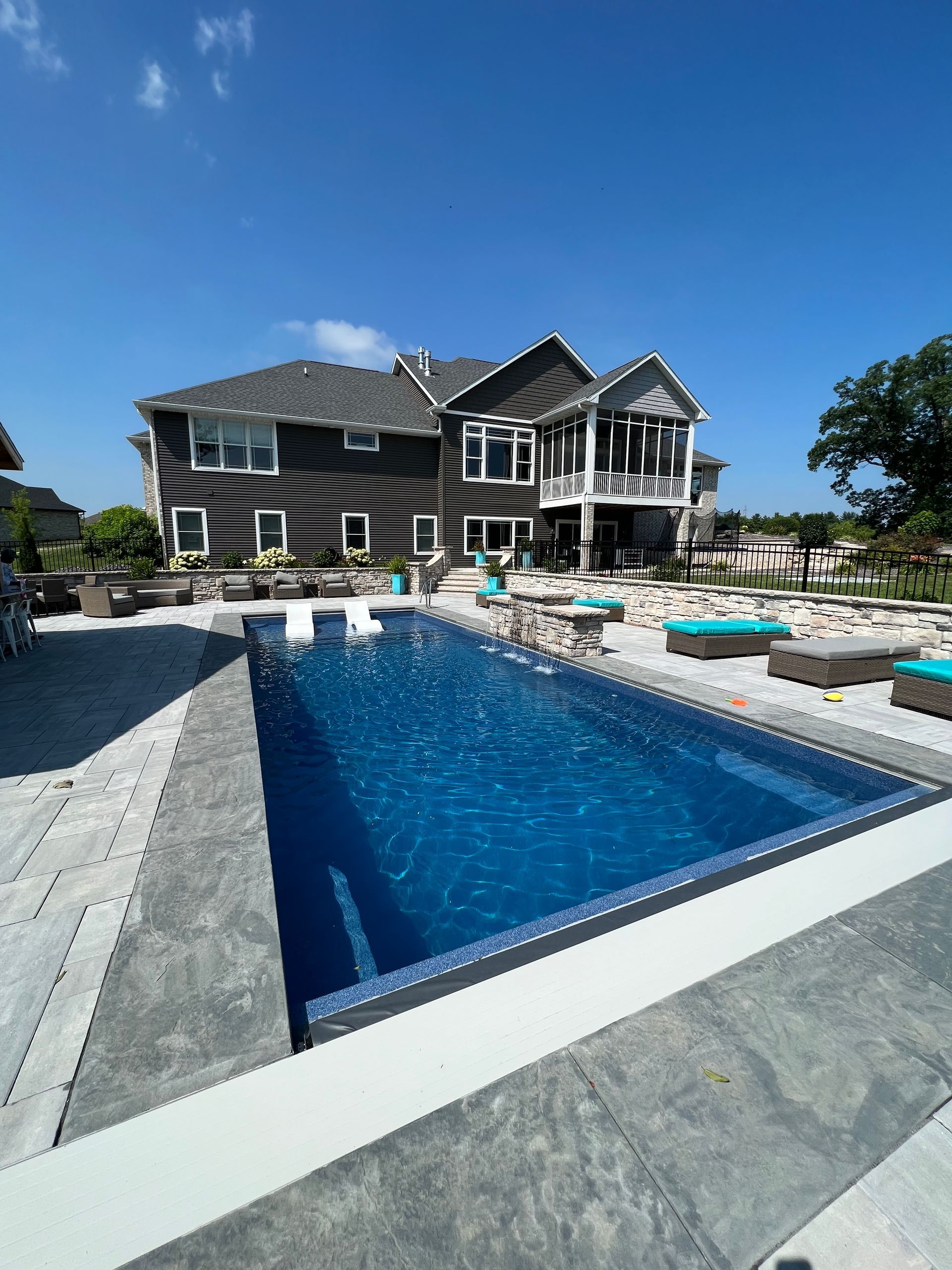 Luxury home with pool; dark gray exterior, blue pool water, patio furniture, and clear blue sky.