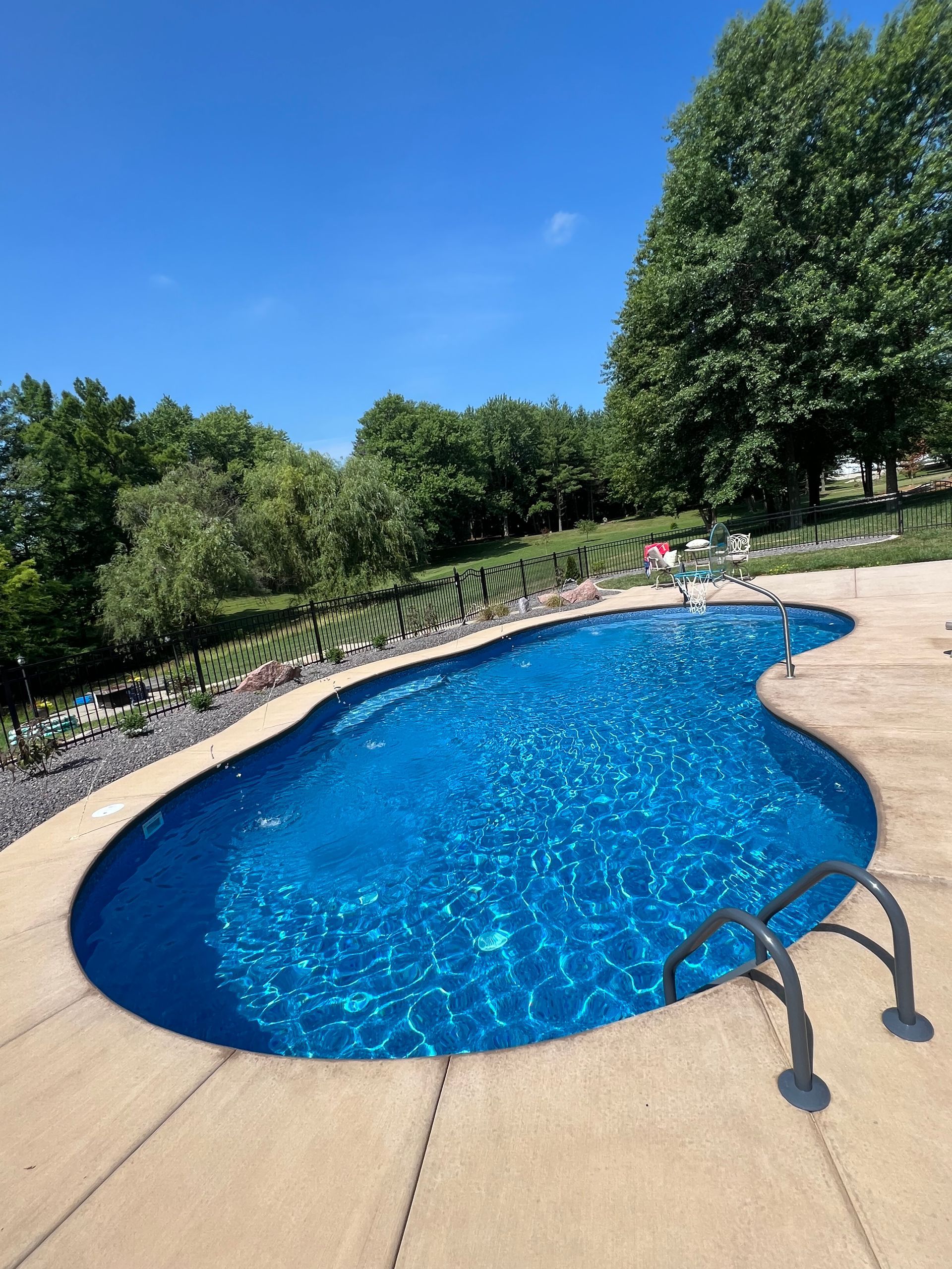 Pool with blue water surrounded by concrete patio, with green trees and blue sky background.