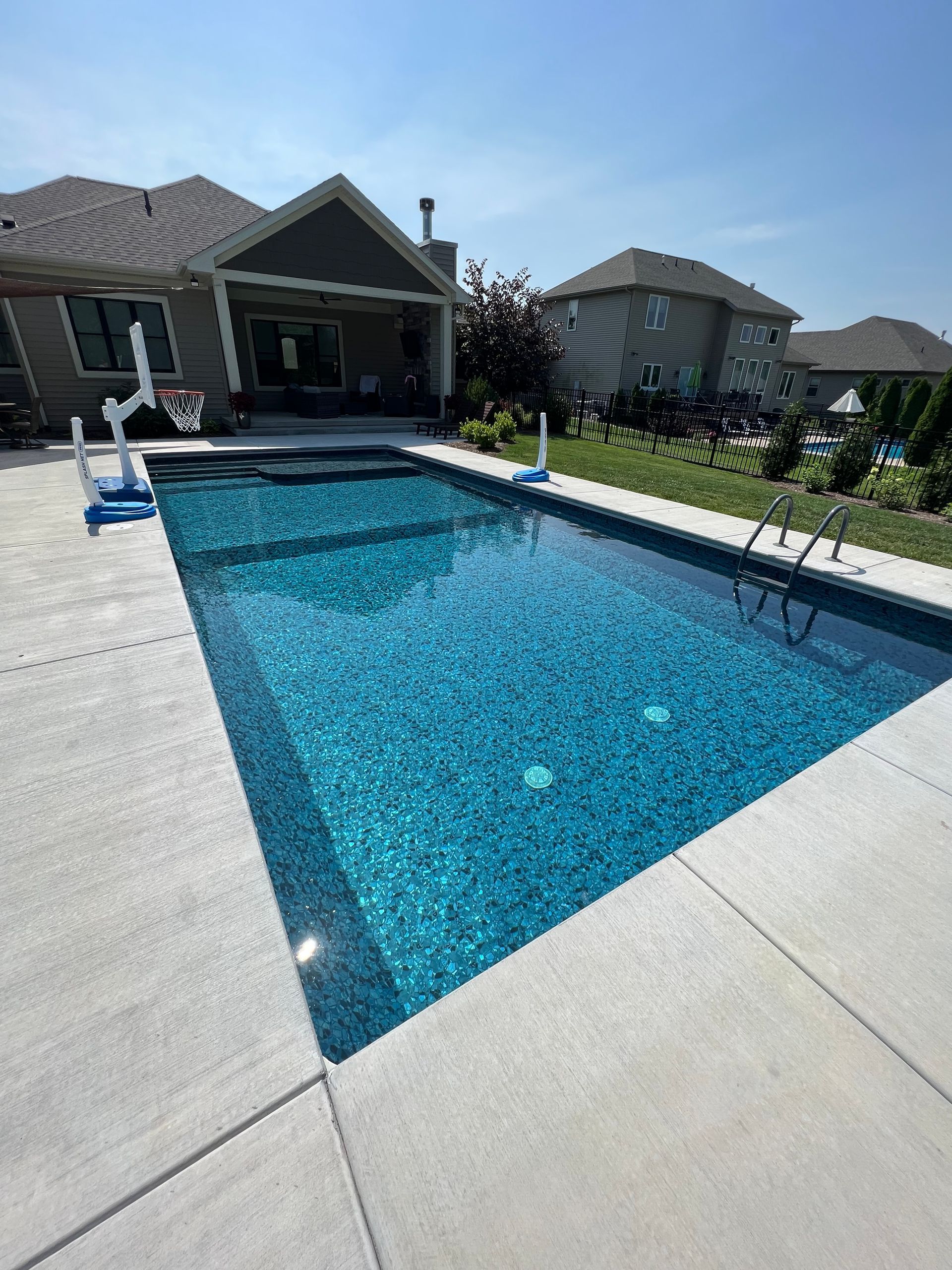 Rectangular turquoise pool surrounded by concrete, next to a house under a clear sky.