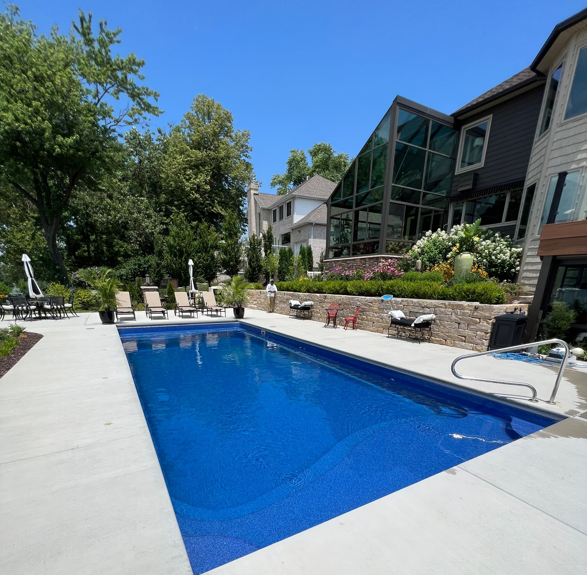 Blue rectangular pool next to a house with outdoor seating and a glass wall, under a clear blue sky.