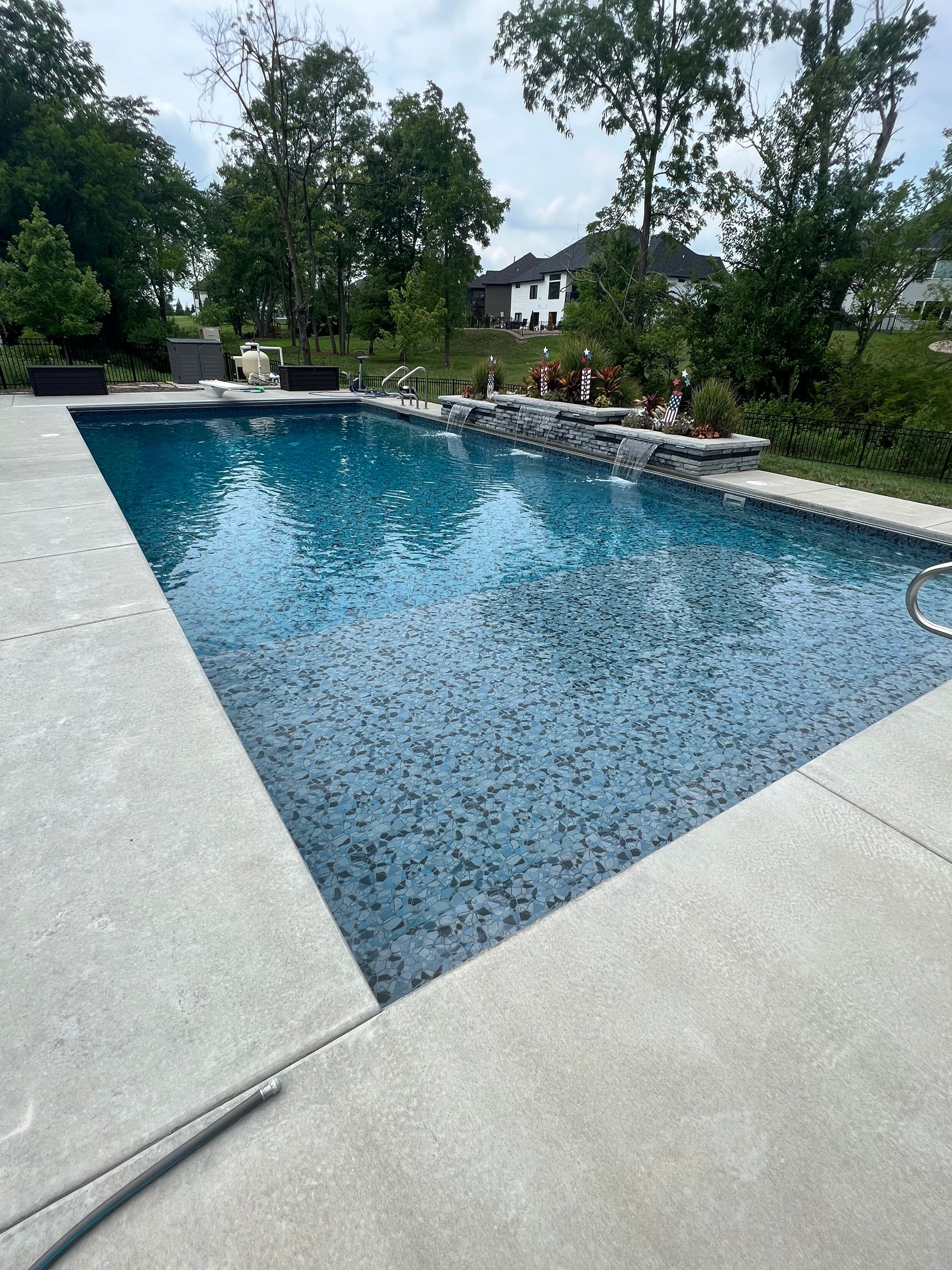 Rectangular swimming pool with blue tile interior, surrounded by concrete. Trees and houses in the background.