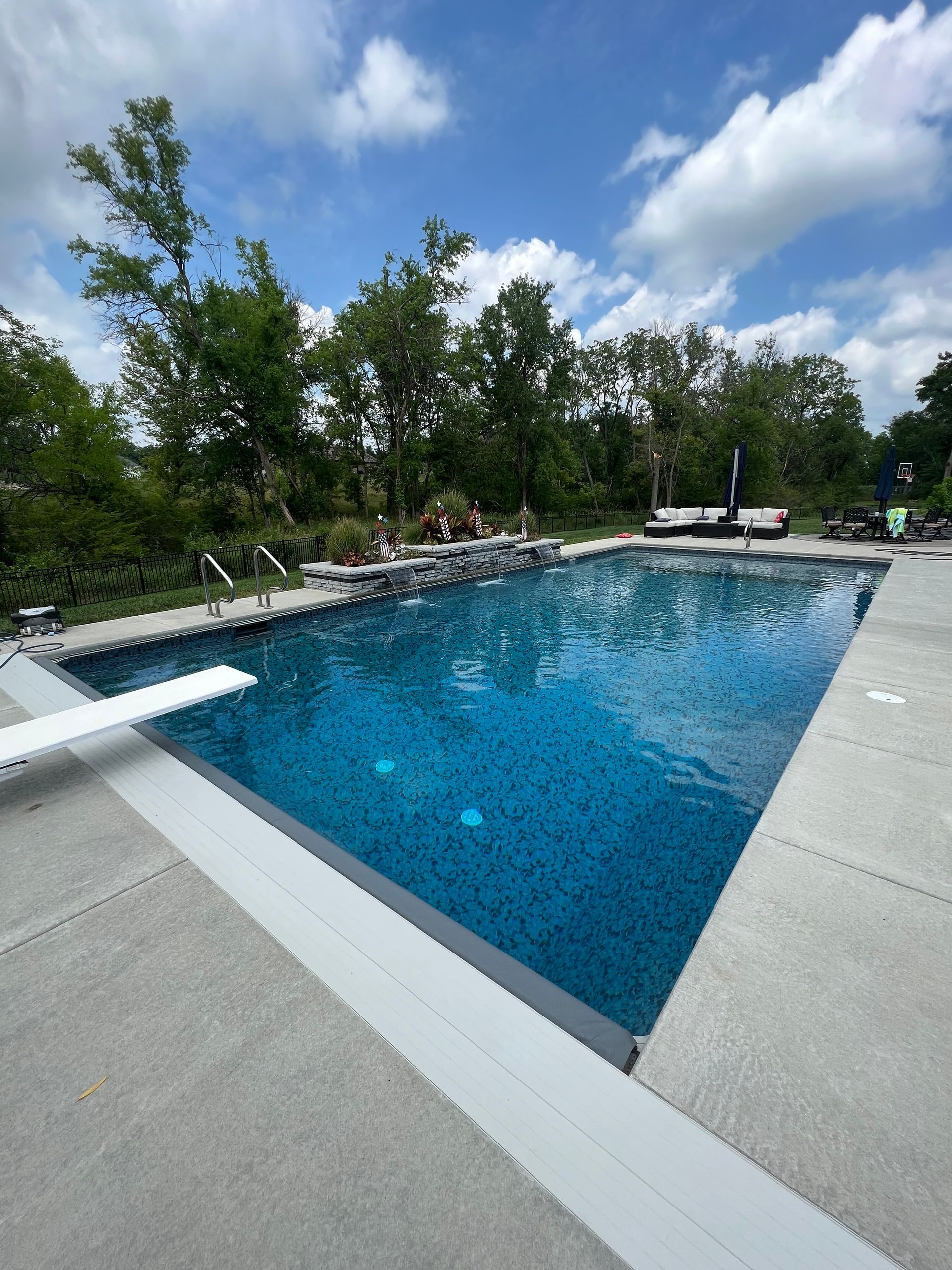 Swimming pool with blue water and diving board; trees and sky in the background.