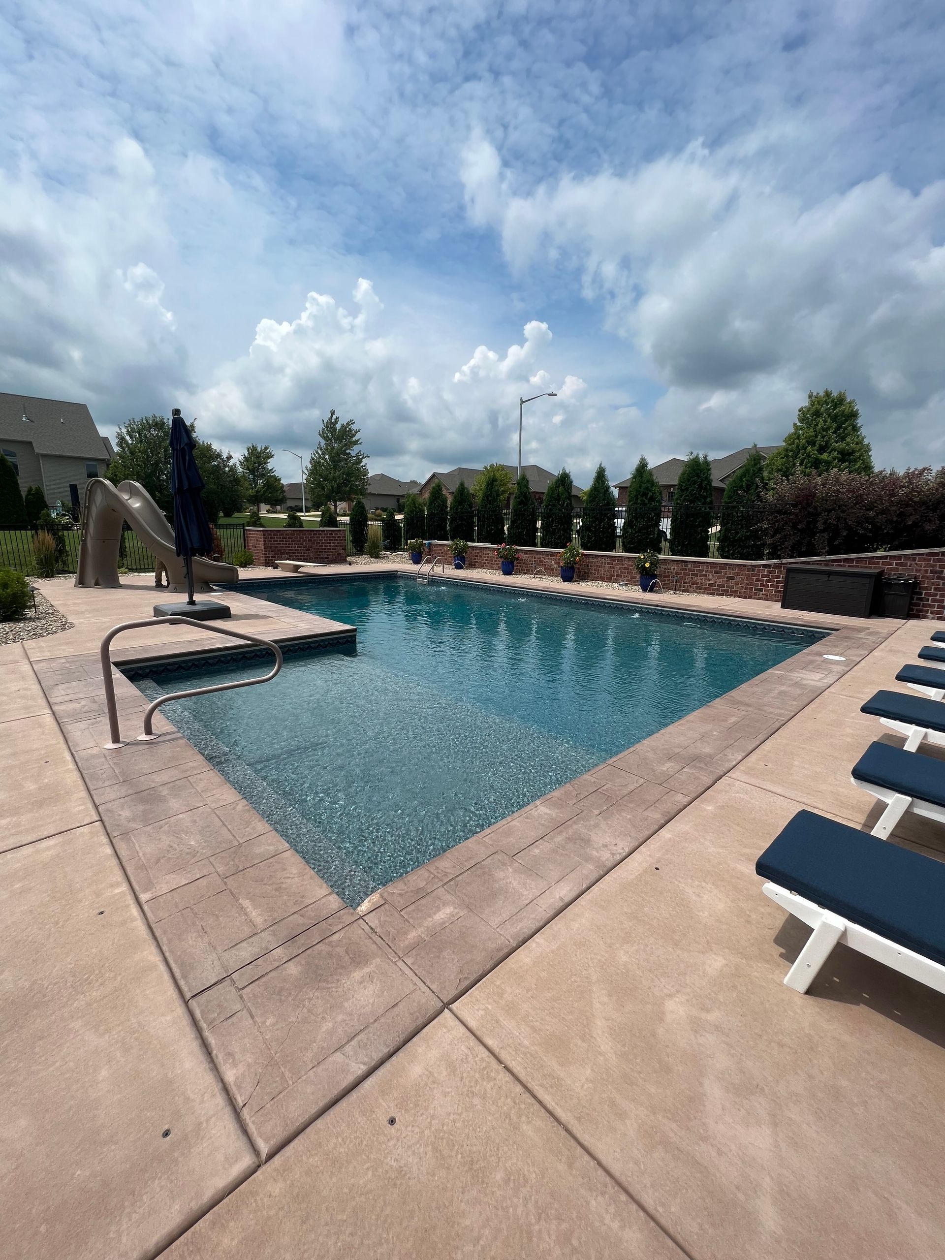 Swimming pool with blue water surrounded by concrete patio and lounge chairs under a cloudy sky.