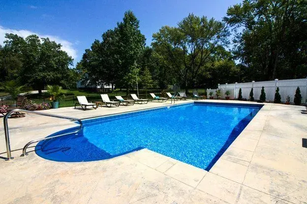 Blue rectangular pool with lounge chairs on concrete patio, surrounded by trees and a white fence.
