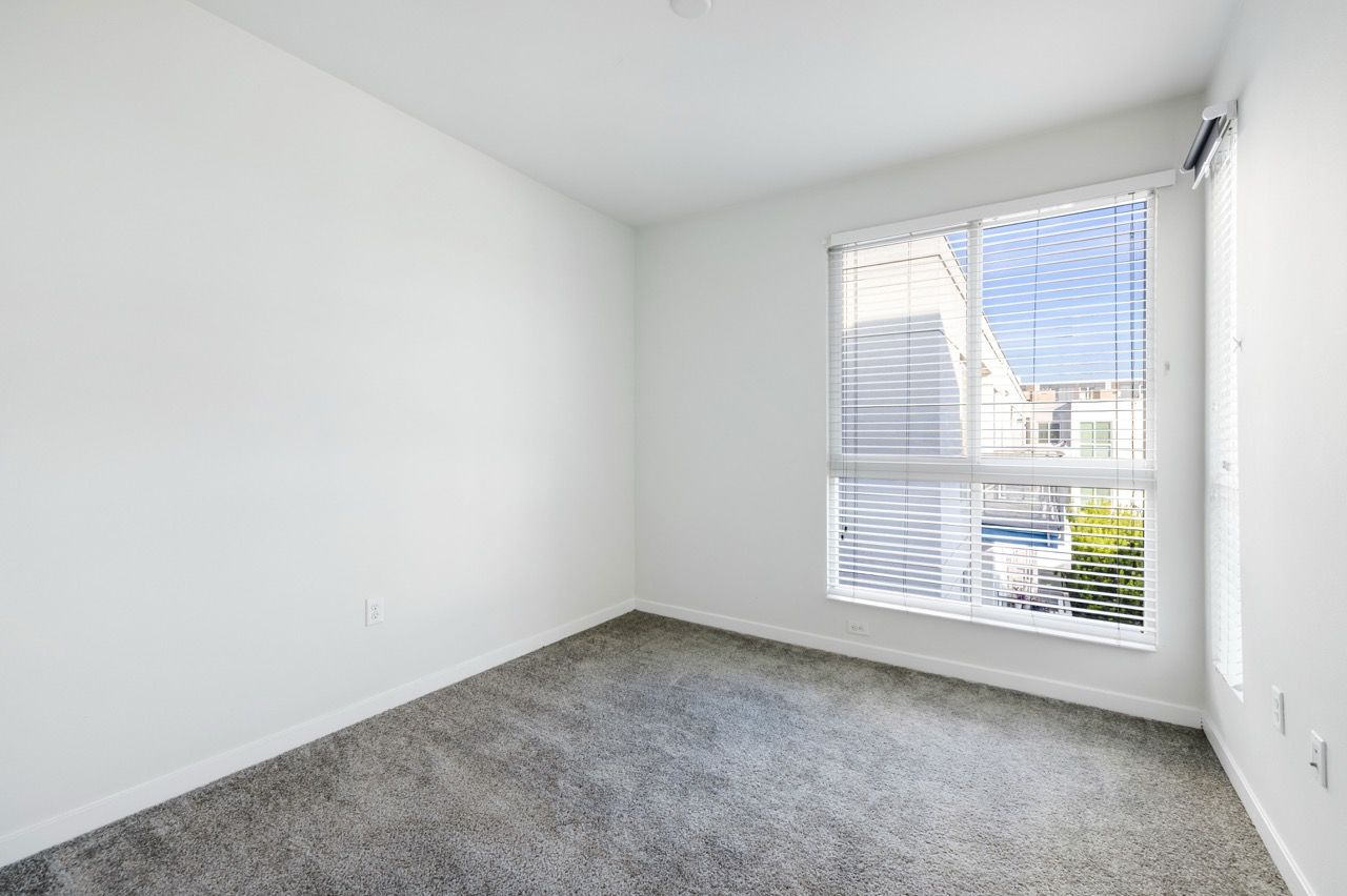 Empty bedroom with white walls, gray carpet, and a large window with horizontal blinds.