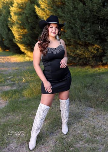 Woman in a black dress and cowboy hat standing outdoors in tall white boots.