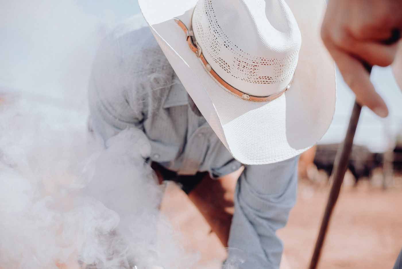 Person in a white cowboy hat crouches in smoke while handling cattle on a ranch
