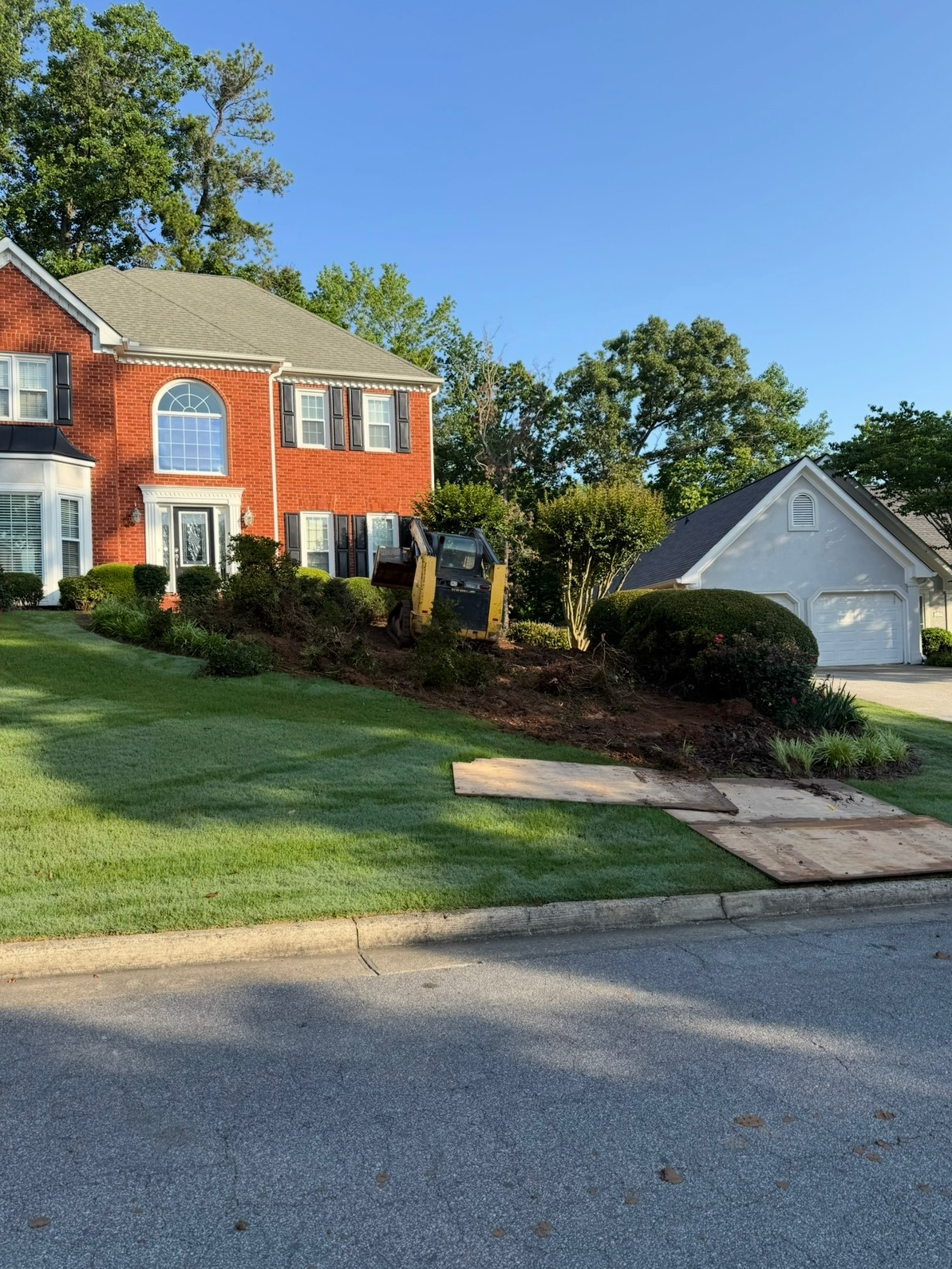 Red brick house with a well-manicured lawn, landscaped beds, and a gray garage on a sunny day.