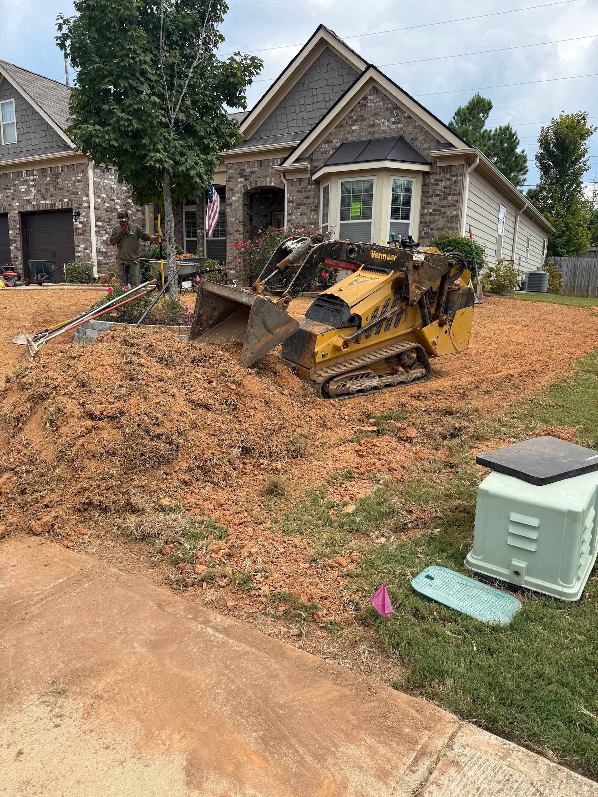 Yellow skid steer operating on a lawn, next to a house with brown brick and a manicured yard.