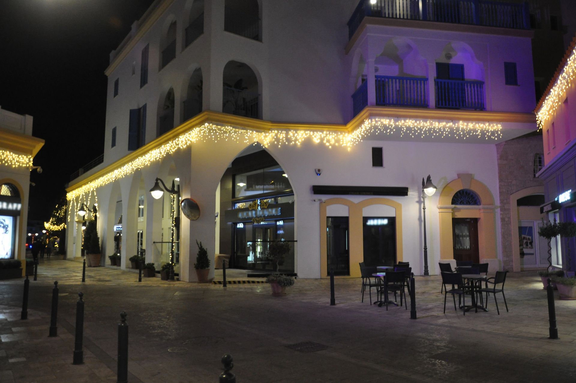 Street at night, white buildings with archways, lit with yellow lights. Tables and chairs outside.