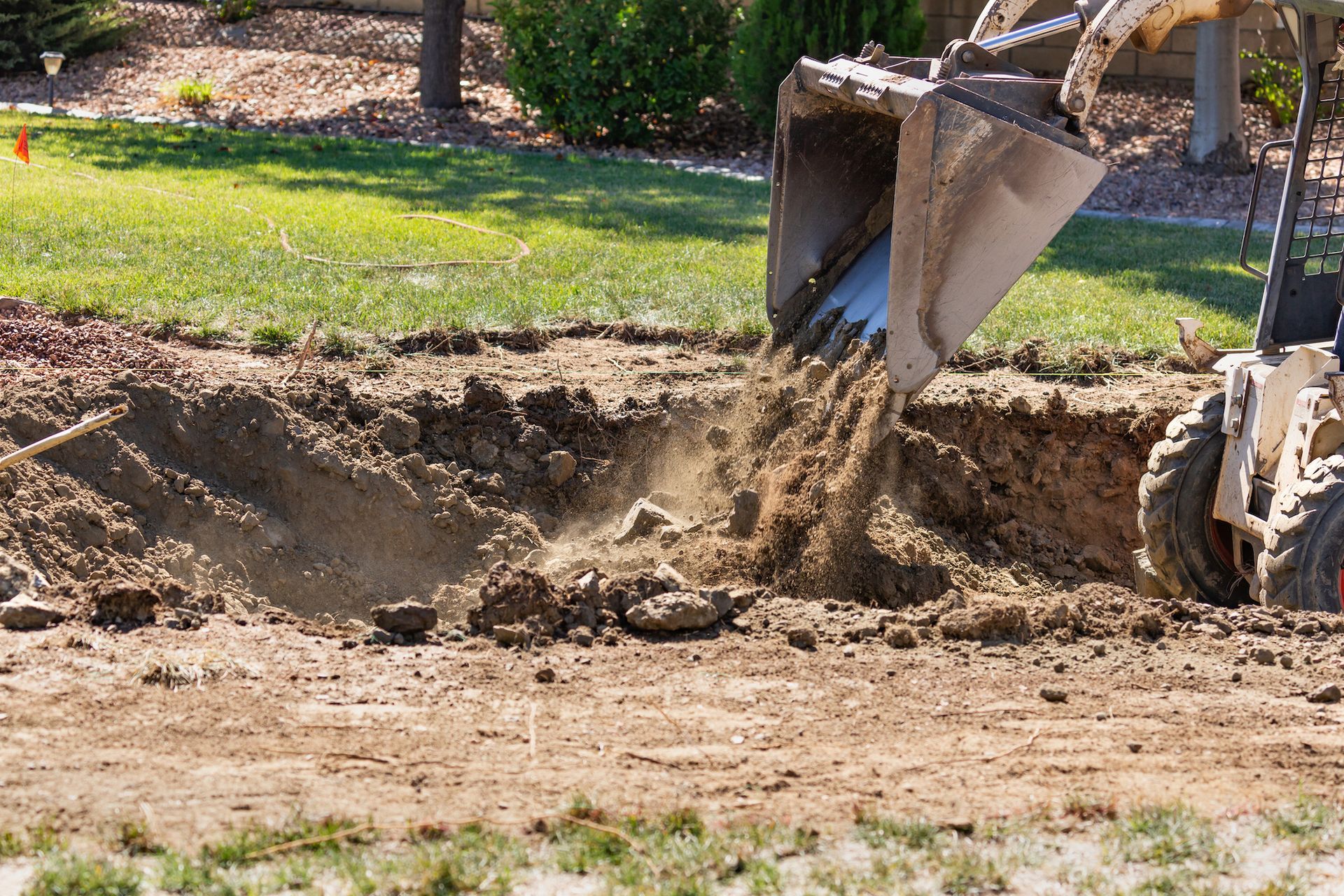 A small excavator dumping dirt into a trench in a grassy yard on a sunny day.