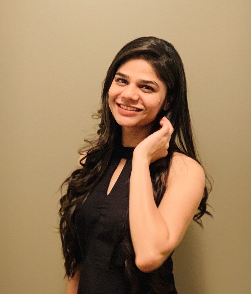 Woman with long, wavy dark hair, wearing a black dress, smiling and touching her hair, posing in front of a neutral wall.