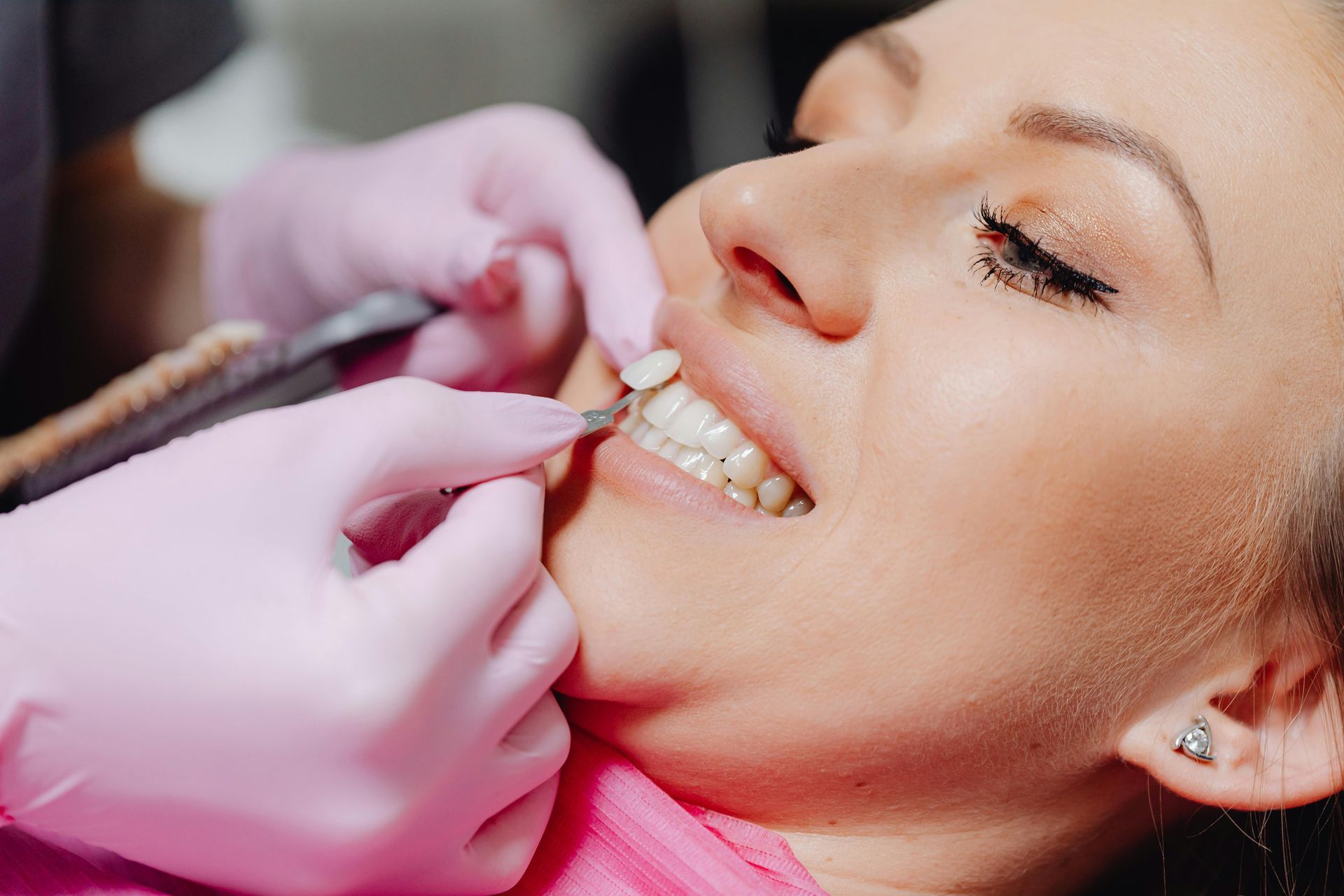 Woman at dentist, pink-gloved hands holding a tool near her teeth, assessing their color.