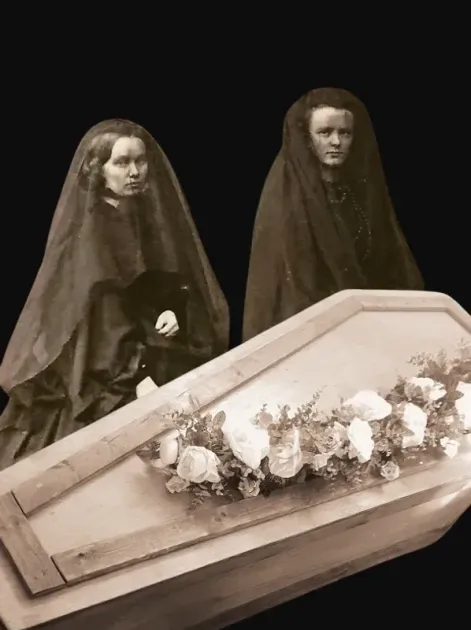 Two women standing next to a coffin with flowers on it