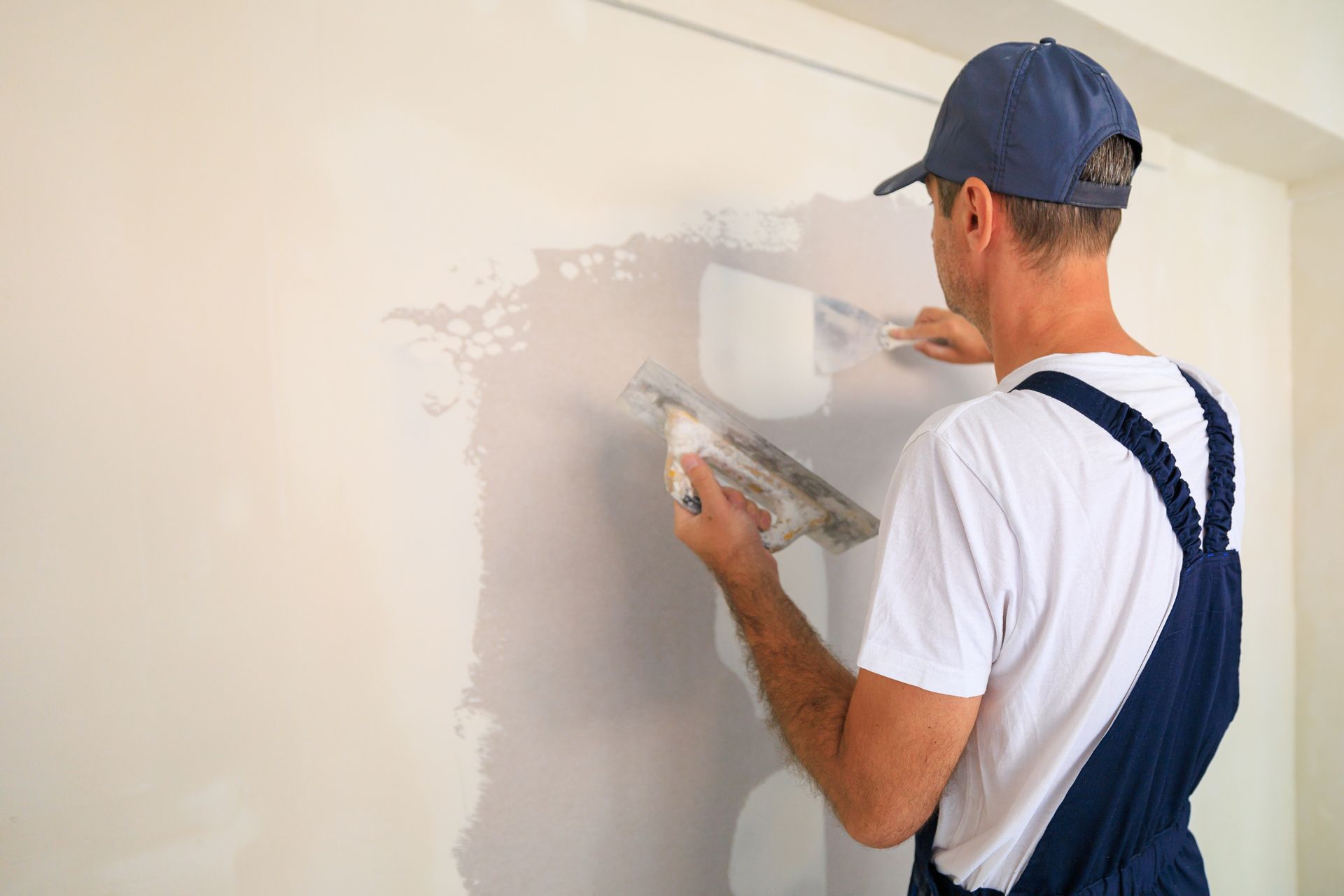 A person in blue overalls and a cap applies plaster to a wall with a trowel and roller.