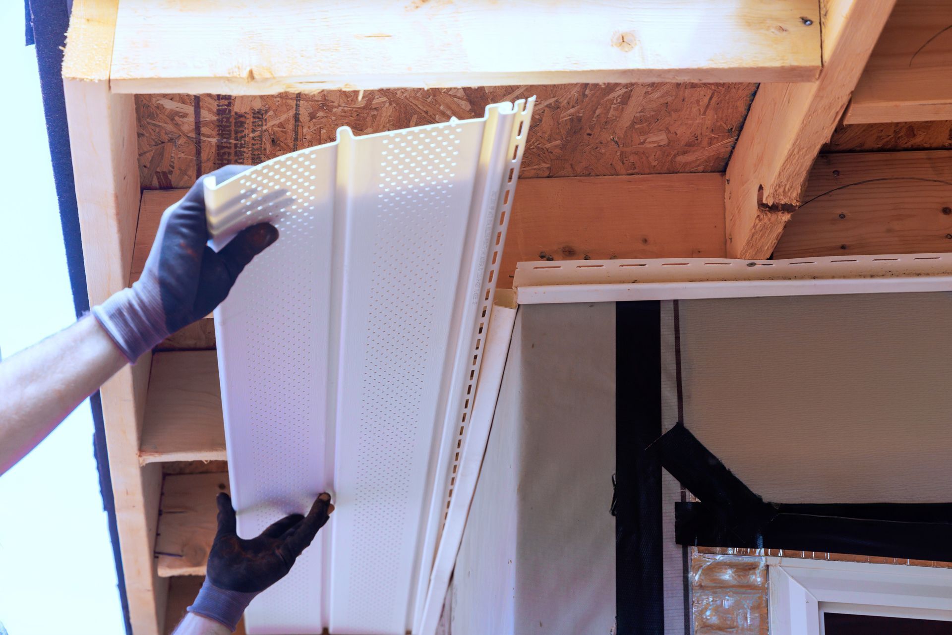 Person installing white vinyl siding on a building, wearing gloves.