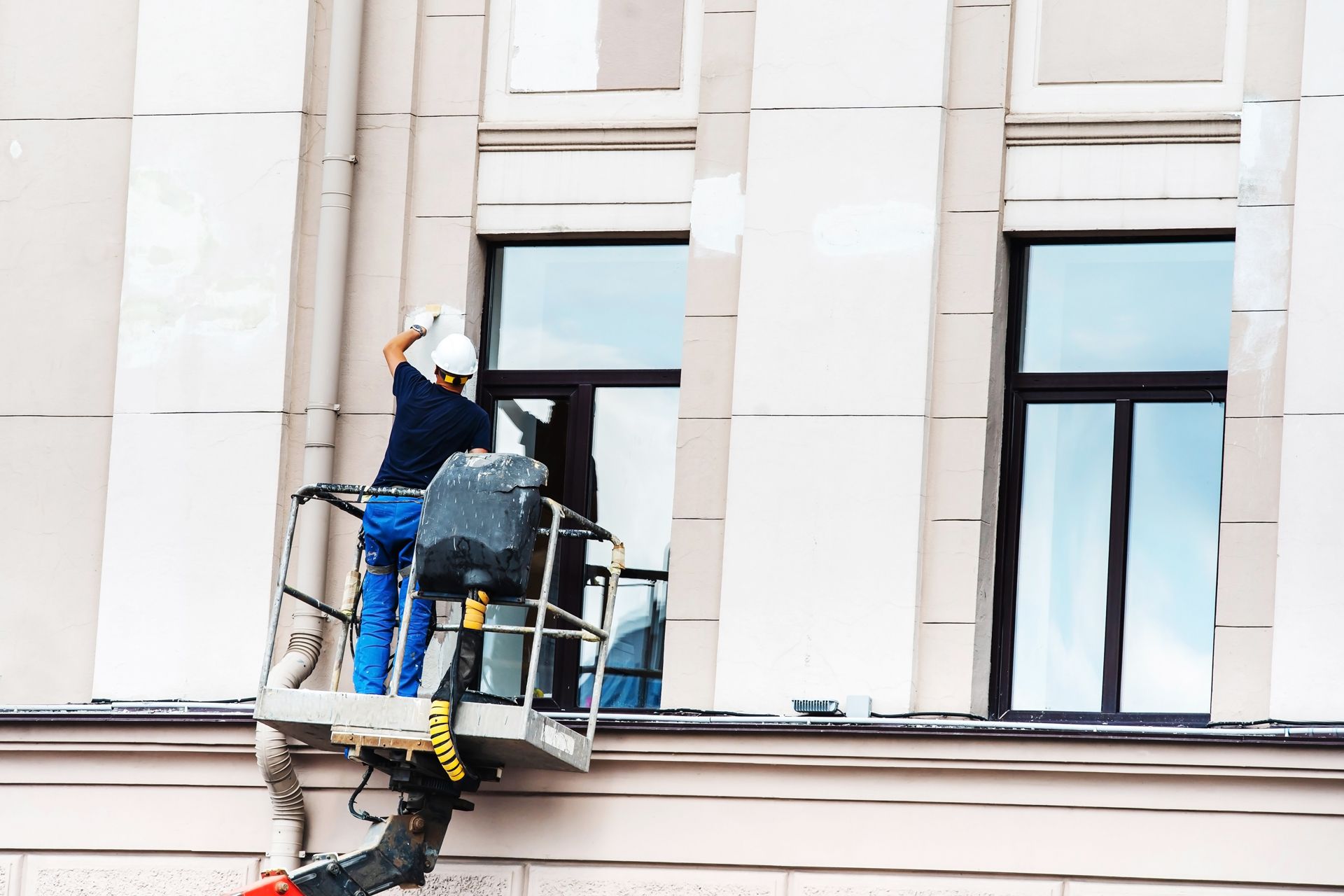 Person in a lift painting a building's facade, windows visible.