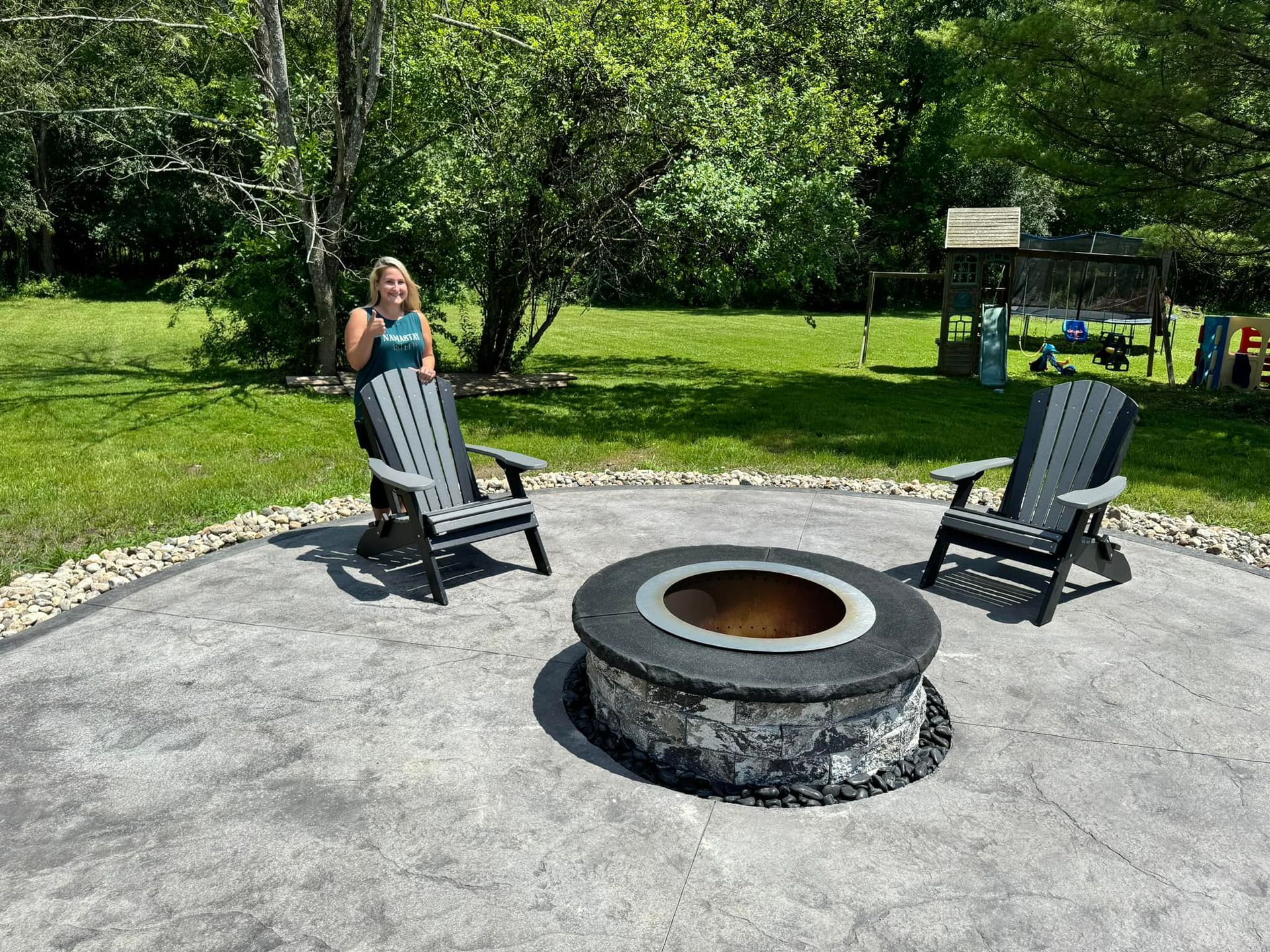 A woman is standing next to a fire pit surrounded by chairs.