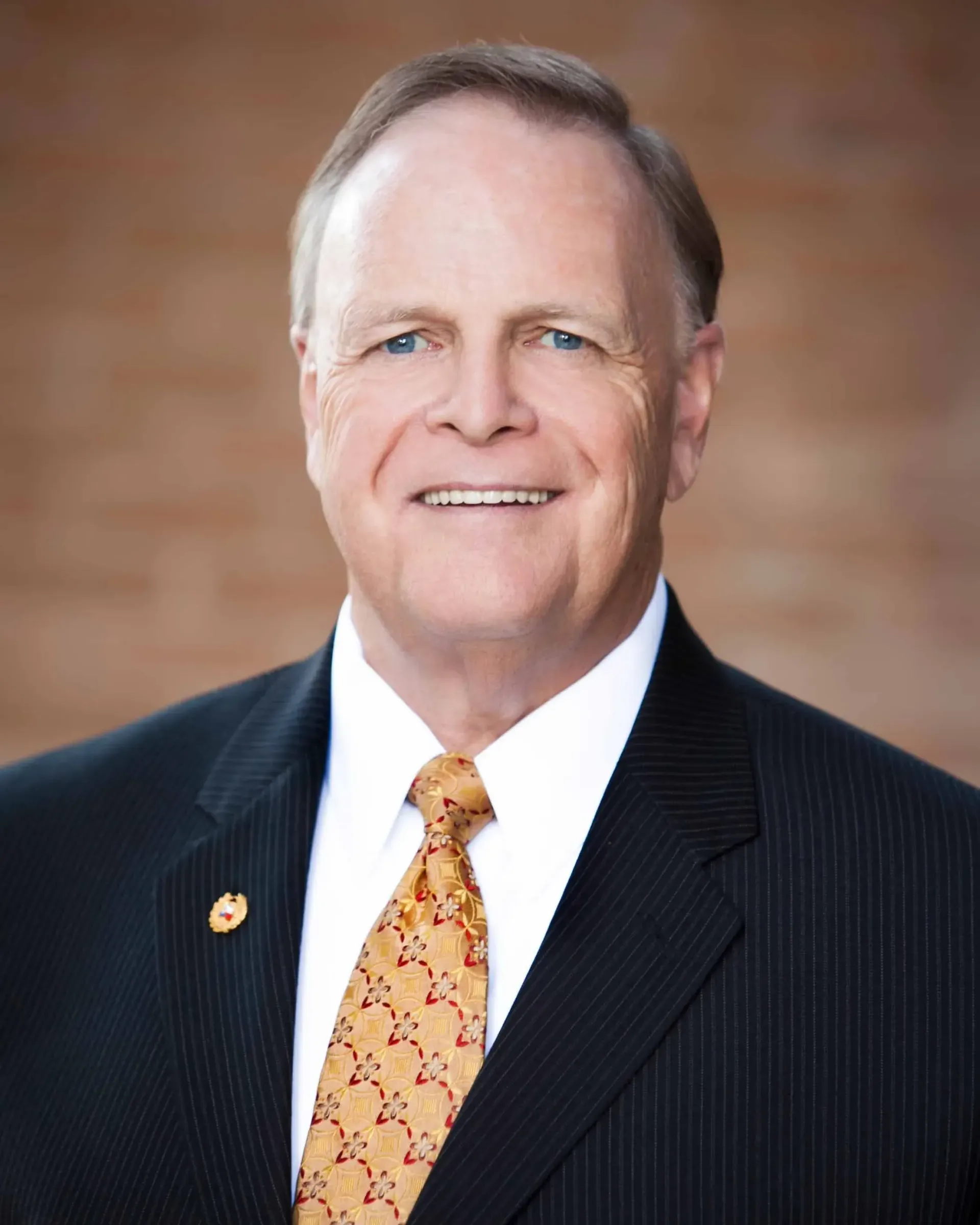 Smiling, older white man in a suit and orange tie, against a brick background.