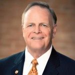 Smiling, older white man in a suit and orange tie, against a brick background.