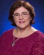 Woman with glasses, smiling, wearing a magenta sweater, necklace, set against a blue background.