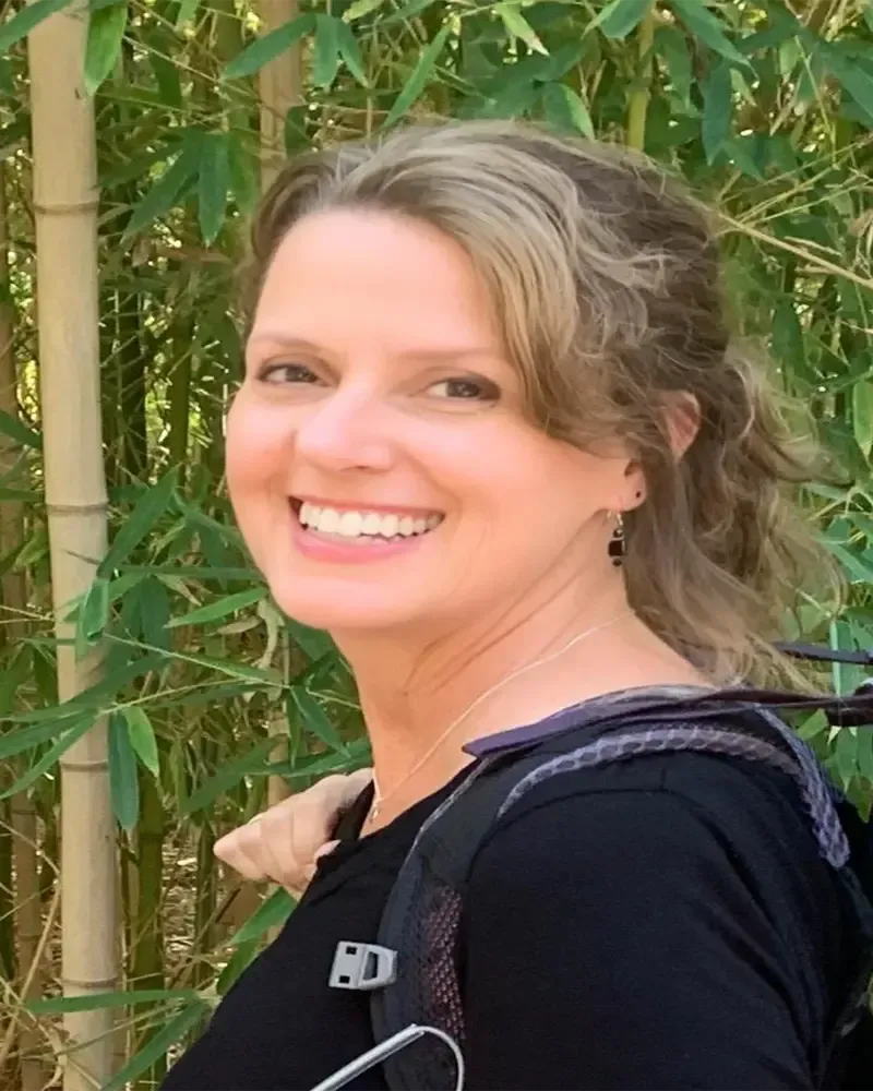 Woman smiles, outdoors. Wearing black shirt and backpack straps. Light skin, blonde hair. Bamboo background.