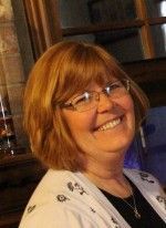 Woman with short, light brown hair and glasses, smiling, wearing a white sweater and black shirt.