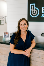 Woman in navy dress smiles, leaning on a reception desk in a modern office.